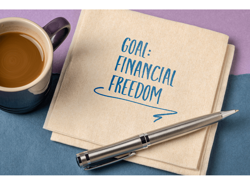 A notepad with "goal: financial freedom" written on it, a silver pen on top, and a coffee mug on a purple and teal background.